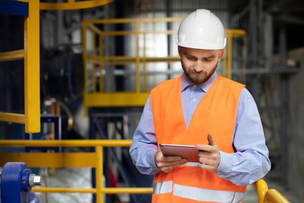 Homem com colete refletivo e capacete usa tablet em ambiente industrial