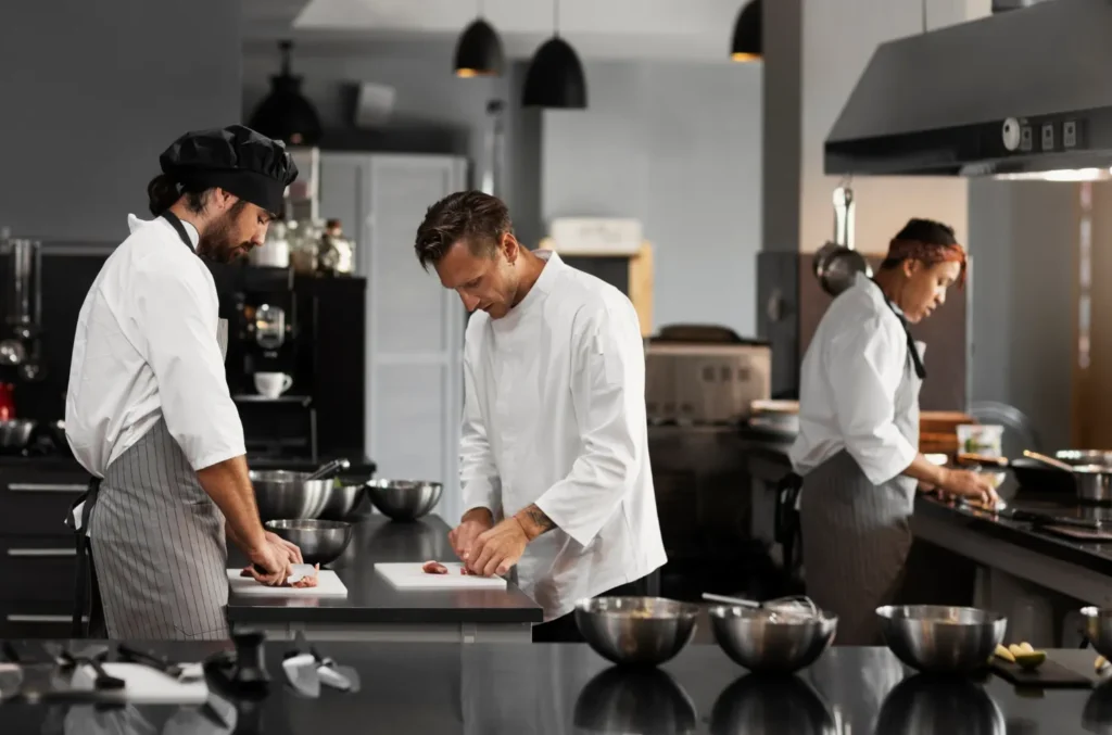 Cozinheiros em pé em uma cozinha de restaurante preparada para produção culinária, com panelas e bancadas de aço inox e ambiente profissional ao fundo.