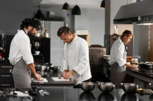 Cozinheiros em pé em uma cozinha de restaurante preparada para produção culinária, com panelas e bancadas de aço inox e ambiente profissional ao fundo.