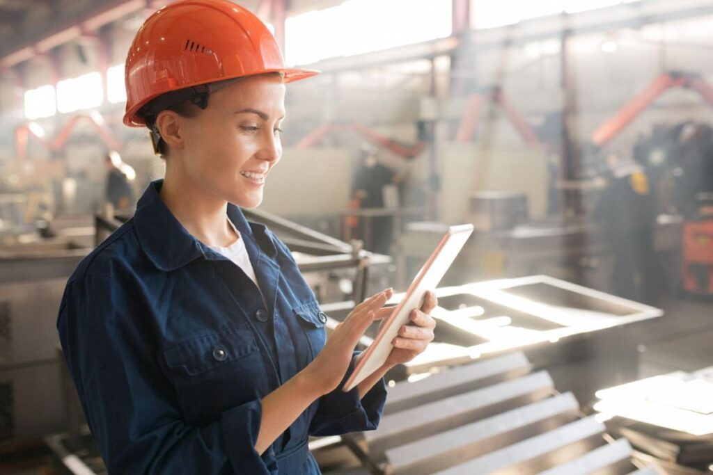 Operária industrial com tablet em mãos e em interior de indústria com maquinários e operadores ao fundo representando o controle de produção