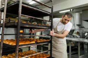Confeiteiro em cozinha de confeitaria assando pães em forno industrial, observando bandejas com dezenas de itens dourados em estantes metálicas.