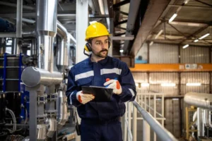Técnico de manutenção em indústria, usando capacete e luvas, segura uma prancheta enquanto monitora equipamentos em uma planta de processamento, com foco em eficiência e economia com GLP.