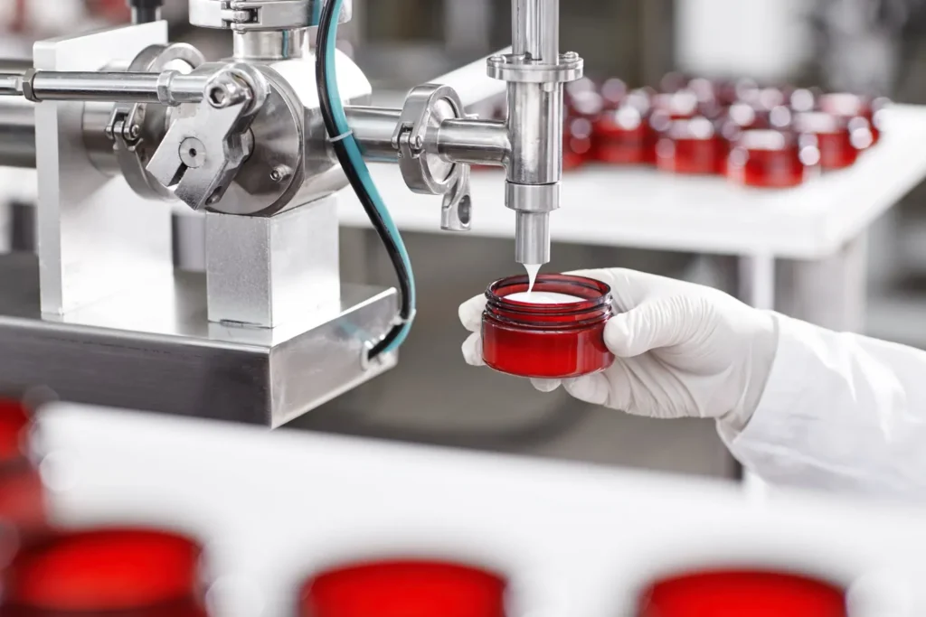 Produção de cosméticos em laboratório: equipamento industrial em aço inox faz o enchimento de frascos com líquido vermelho, enquanto um profissional com luvas conecta o sistema e controla o processo.