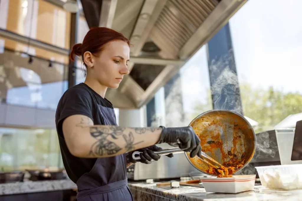 Trabalhadora com luvas cozinha em food truck com chama e fumaça saindo de panela, demonstrando preparo durante operação. Instalação de gás em food truck